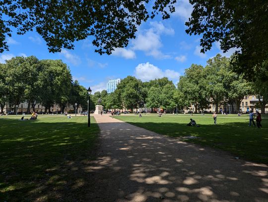 A photo of a square in the summer in a city. A path leads through the centre to a statue of a man on a horse. The square is lined by big green trees.